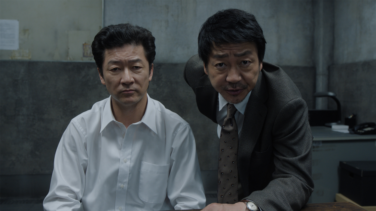 薄暗い取調室の中で白いシャツを着て、リーゼントの男(浅野忠信)とグレーのスーツを着た男(大森南朋)が正面をにらみつけている