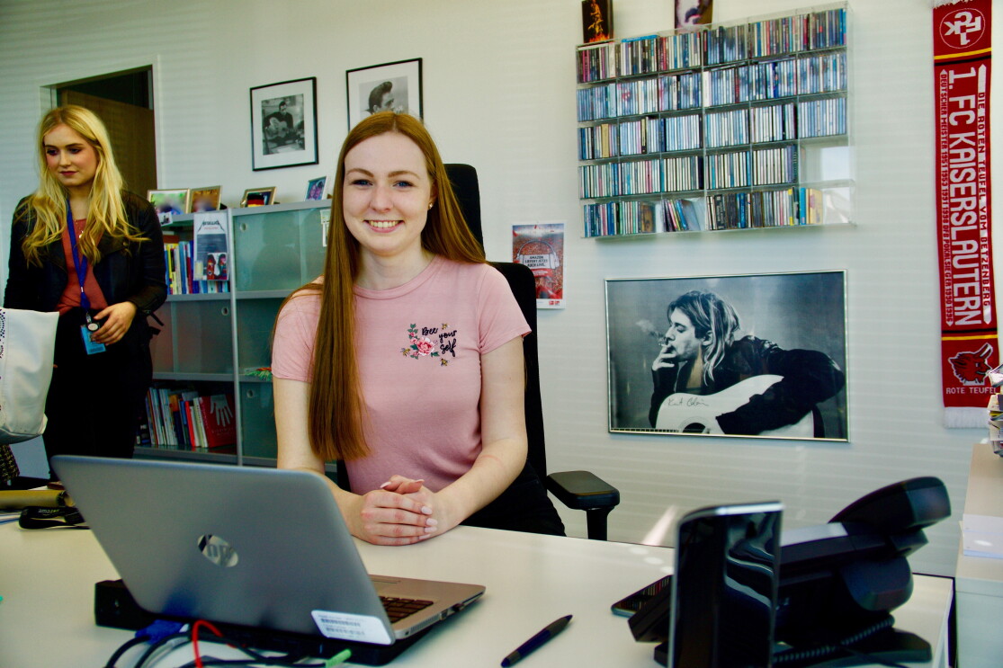 Eine junge Frau in rosa T-Shirt sitzt an einem Schreibtisch, vor ihr ein Laptop