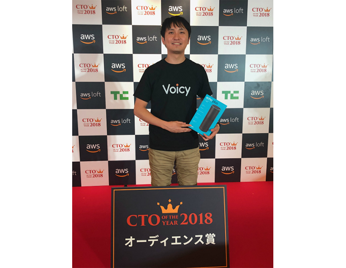 白と黒の市松模様の壁を背に立つ男性。黒いTシャツで手に水色の箱を持っている。足元に黒いボード、床は赤。