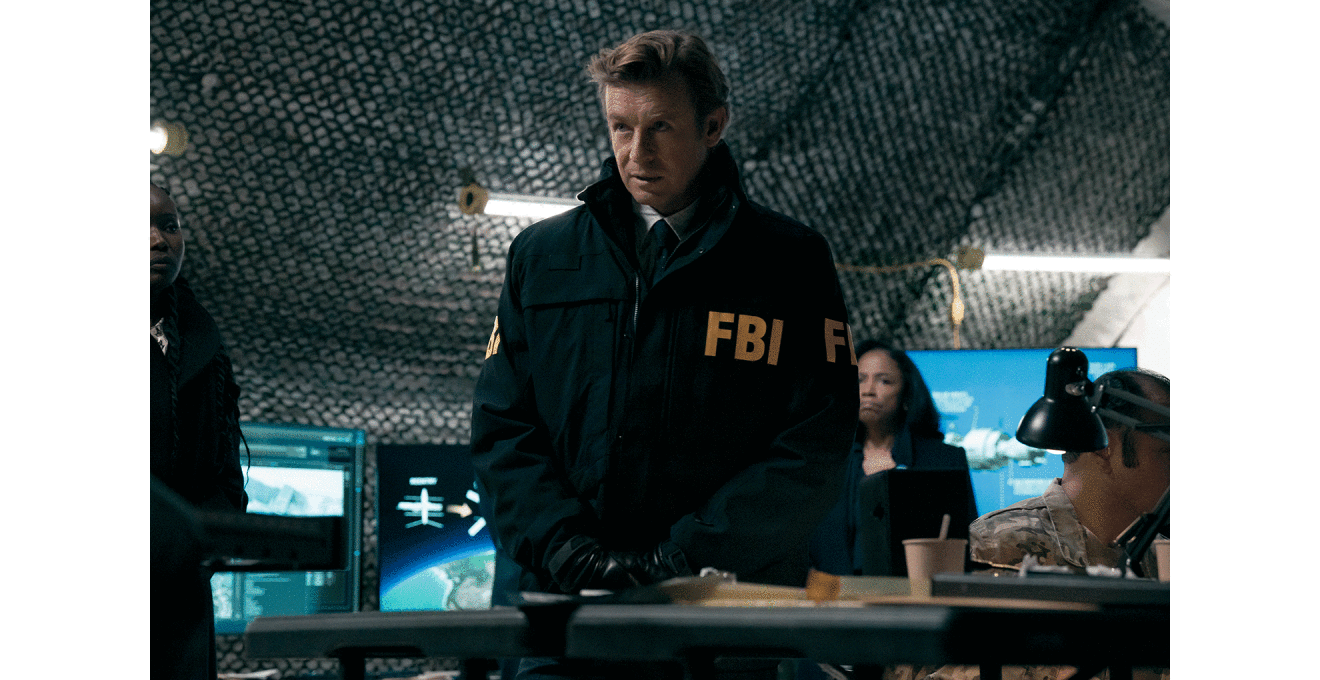 薄暗い室内で、FBIの文字が入った濃い色のジャケットを着た男性が立っている。男性の背後には、大きなモニターが複数並んでいる。