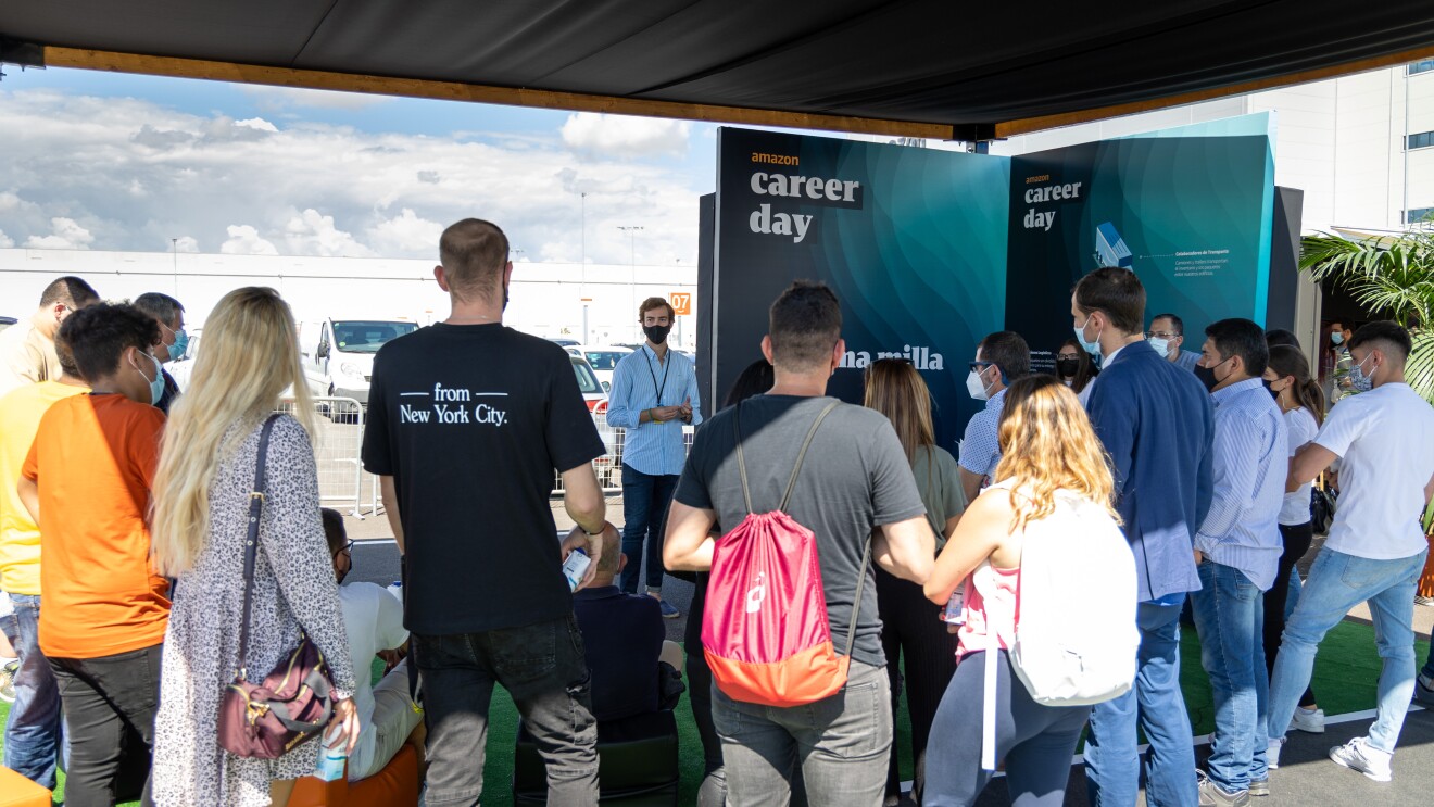 En España, el Career Day contó con una parte virtual y otra presencial, que se celebró en el nuevo centro logístico de Amazon en Illescas (Toledo). En primer plano un grupo de 20 personas de pie y se ve también que hay gente sentada en unos sillones de color naranja. Todas ellas de espaldas. Les habla una persona que está de pie y que se ve gesticular con mascarilla. De fondo el centro logístico de Amazon en Illescas. La gente está escuchando y en la parte derecha hay un cartel que llega al suelo y pone Amazon Career Day y Úlitma milla. Se ve el cielo azul con nubes blancas.