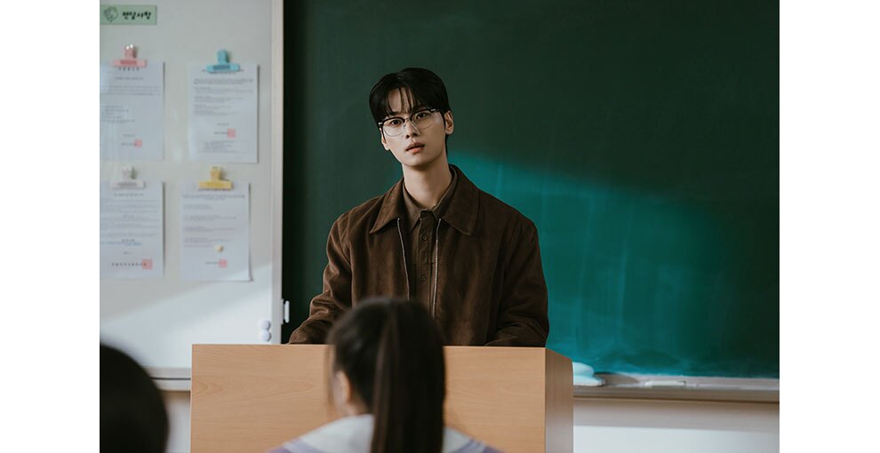 黒板の前、教壇に向かって立つ眼鏡の男性。教壇の手前に生徒2人の後頭部。