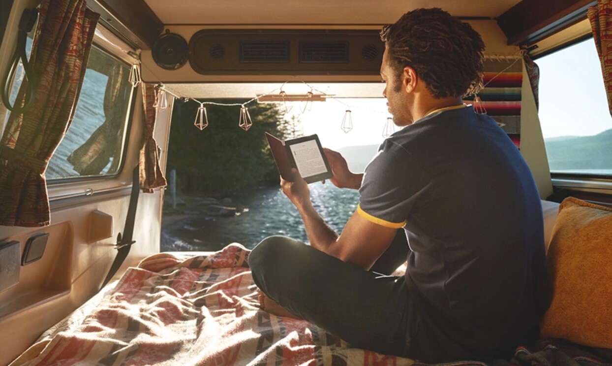 A man sits in the back of a camper van, in front of a large body of water. He is holding a Kindle Paperwhite and appears to be reading.