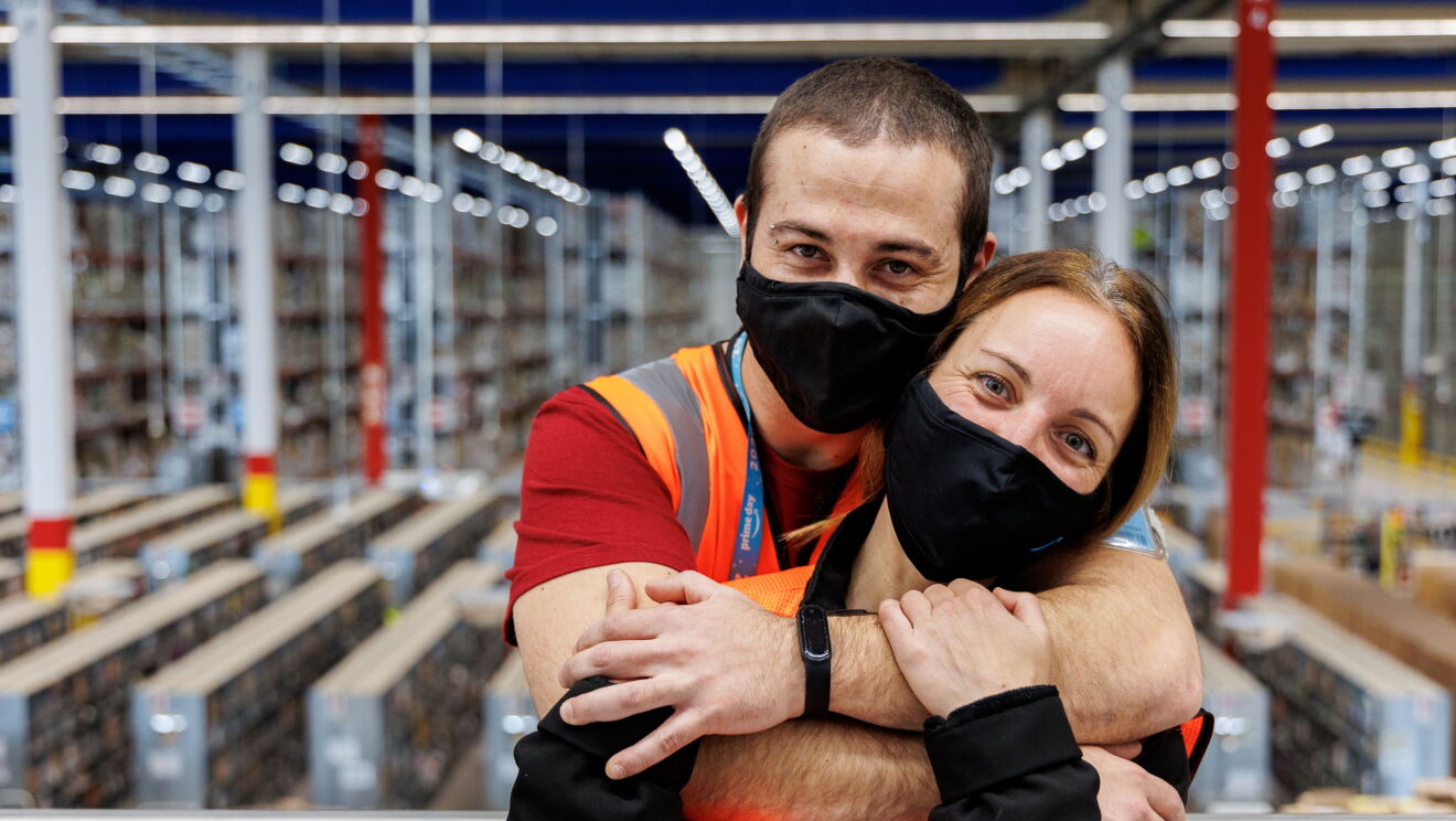 Sira Valeriano y Cristian Gamella. Foto de medio plano mirando a cámara con mascarilla de color negro con la sonrisa azul de Amazon Es una foto interior, en el centro logístico de Illescas que es donde trabajan juntos. Los dos protagonistas miran a cámara abrazados. Cristian, por detrás, abraza a Sira y ella le agrarra una mano y el brazo. Él va con una camiseta roja de manga corta y ella una sudadera y él va con el pelo corto y ella largo recogido con una coleta. Ella tiene los ojos azules y él marrones. 
