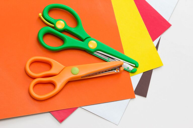 緑色とオレンジ色の安全ハサミが数色の折り紙の上に置かれている