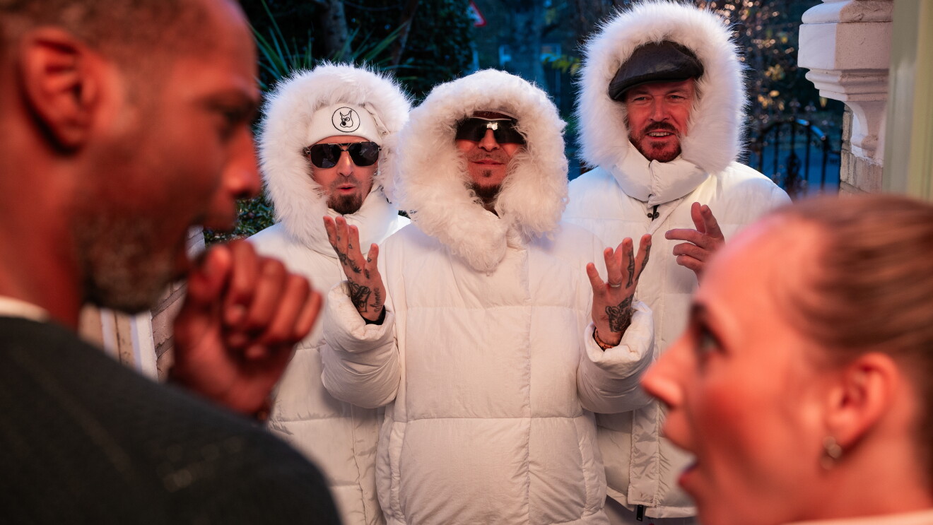 East 17 band members appear in white puffer jackets at a home with a surprised and excited couple. 