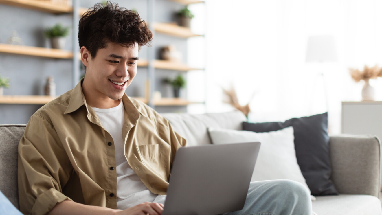 Portrait Of Smiling Man Sitting On The Couch Working On Pc Laptop Indoors In Living Room