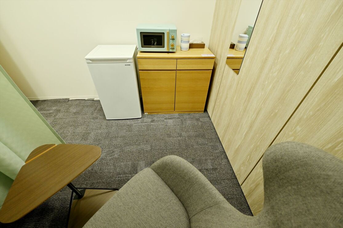 壁に沿って冷蔵庫、棚、電子レンジ、消毒用アルコールが置かれている。その手前に一人用のソファがある