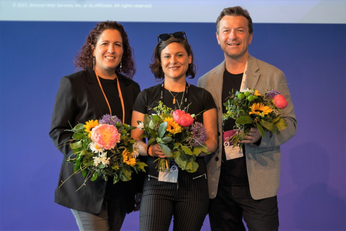 Gruppenbild der AWS-Jury, bestehend aus AWS DACH Chef Klaus Bürg, Softwarearchitektin Nahia Orduna und AWS Community Builder Linda Mohamed, mit Blumensträußen vor einer blauen Wand beim Wettbewerb von AWS GetIT in 2022.