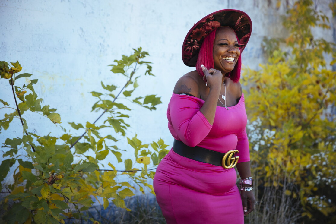 Auhzha Wright smiles as she models her pink dress and custom-made hat while standing outside in front of lush landscaping.