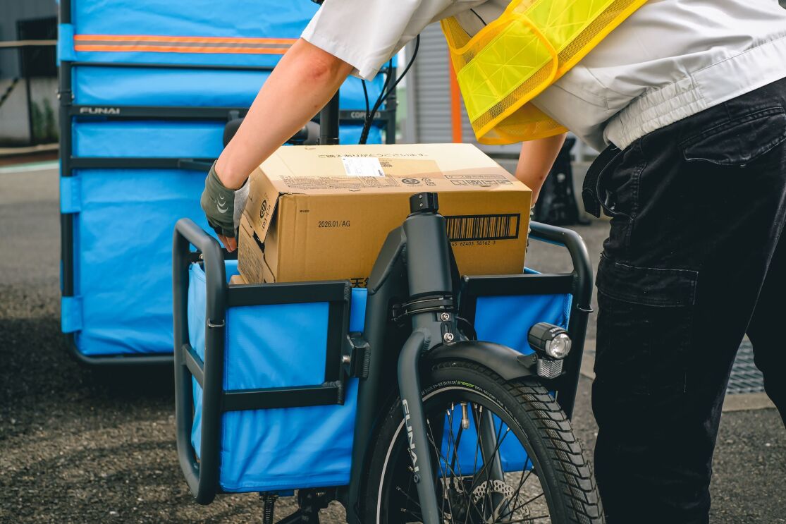 黄色い安全ベストを着ている人が大きなダンボール箱を自転車の前輪部分の青いかごに入れている
