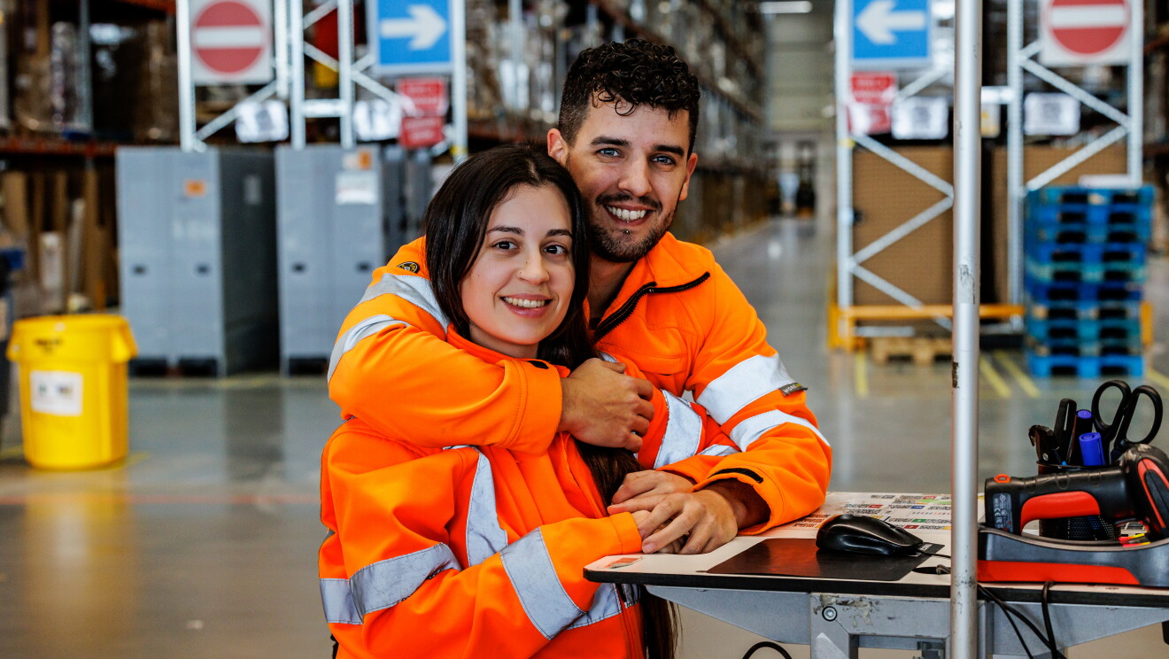 Miriam y Mario posando para la foto mirando a cámara. Él por detrás le abraza y ella le agarra una de las manos. Los dos van con una chaqueta naranja que simula un chaleco de seguridad. De fondo el pasillo de un centro logístico. Miriam lleva el pelo largo por la cintura y es de color castaño. Él tiene el pelo corto pero la parte superior la lleva un poco más larga y la tiene rizada. Tiene los ojos claro, barba de dos días y un piercing en el labio inferior. Están apoyados en una mesa alta en la que se ve un lector de barras y un ratón de ordenador.