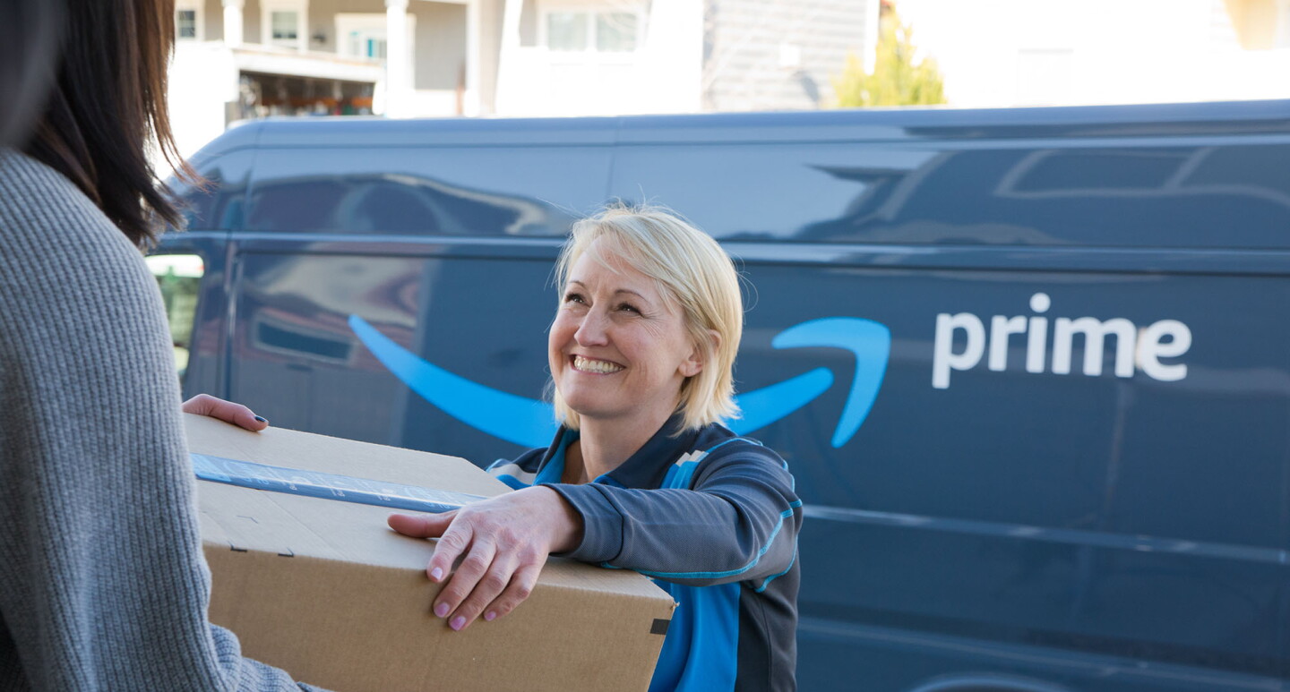 A woman delivers a box to another woman. The delivery driver stands in front of an Amazon DSP van. 