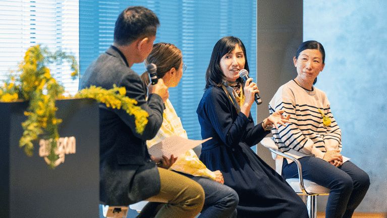 ブラインドがかかった室内で、並んで椅子に腰かける4人の人たち。左端の男性と、右から2番目の女性がマイクを持っている。