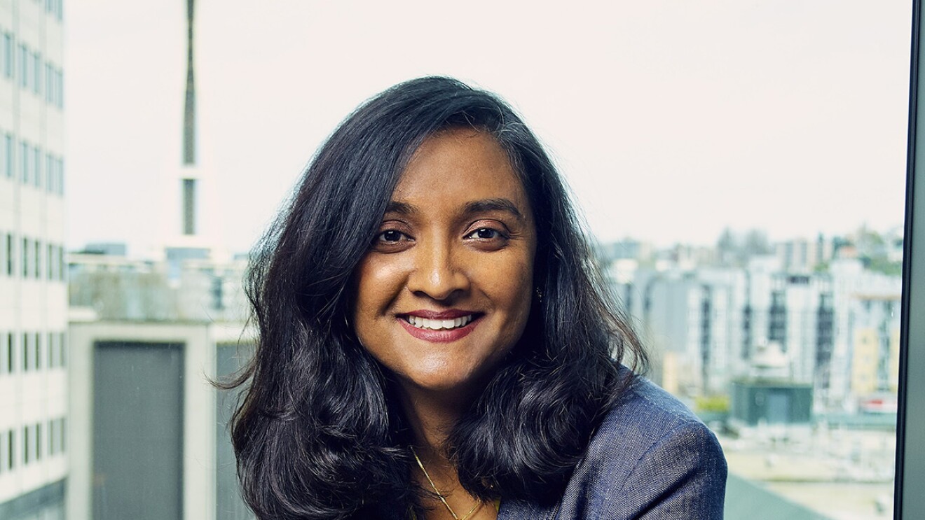 A woman sits on the ledge of window in an Amazon office building. She wears a blazer over a bright yellow top and jeans, smiling at the camera.
