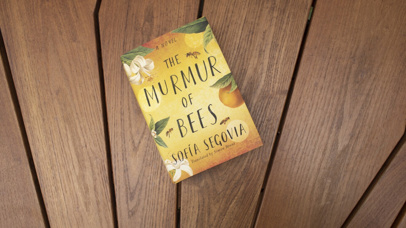 Book cover for "The Murmur of Bees" on a teak wood background.