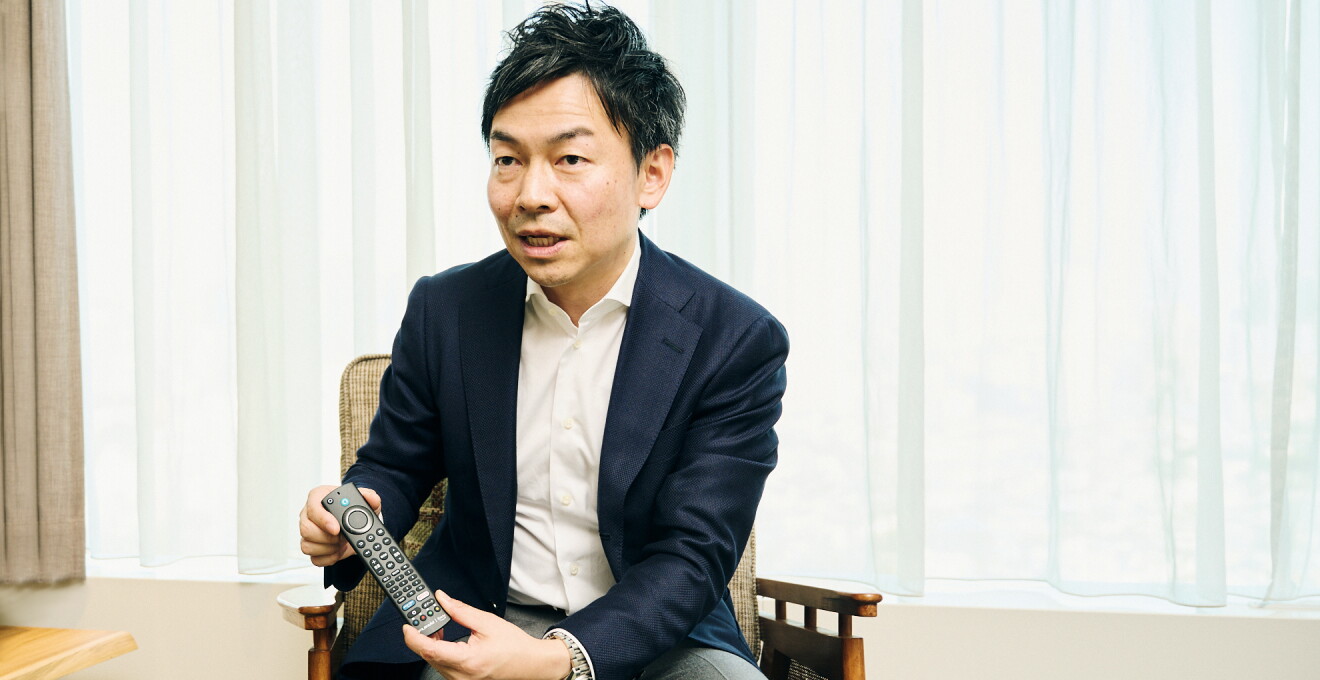 黒い「FUNAI Fire TV搭載スマートテレビ」のリモコンを手に持つAmazonデバイス Fire TV 事業部 事業部長の西端明彦さん