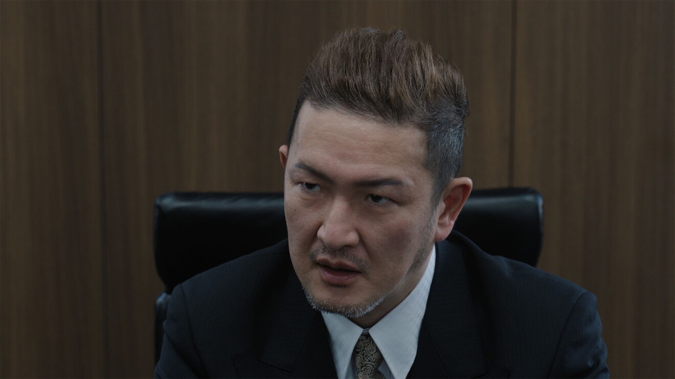 木製の壁の前に置かれた黒い革のハイチェアに座るダークスーツを着た茶髪の男(中村獅童)が険しい表情で話している
