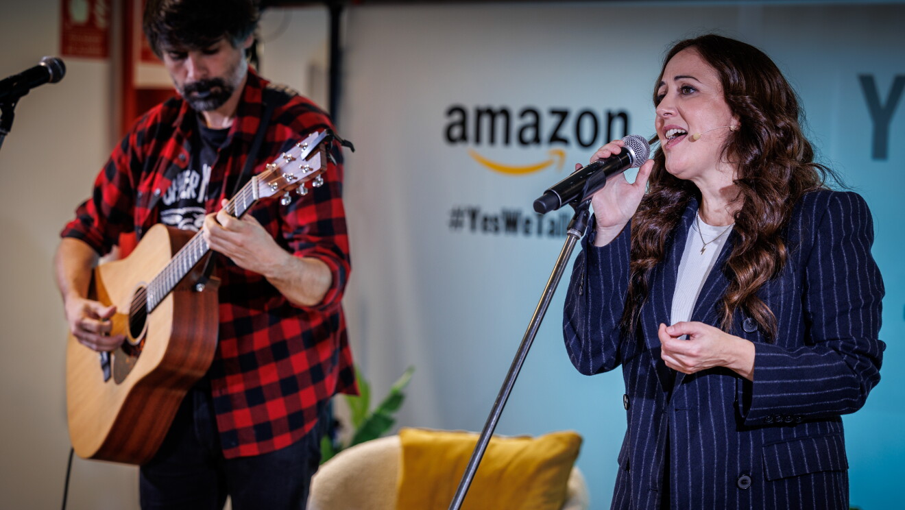 En un escenario creado para la ocasión, de fondo hay un cartel con el logo de Amazon y el Yes, we talk, que es el lema de la campaña. En primer plano la cantante Mai Menses con una americana azul de rayas y un jersey de punto blanco agarra con una mano el micro mientras canta. En segundo plano y desenfocado el guitarrista con una guitarra española, una camisa de cuadros negra y roja, barba negra. Lleva una púa en la boca. 