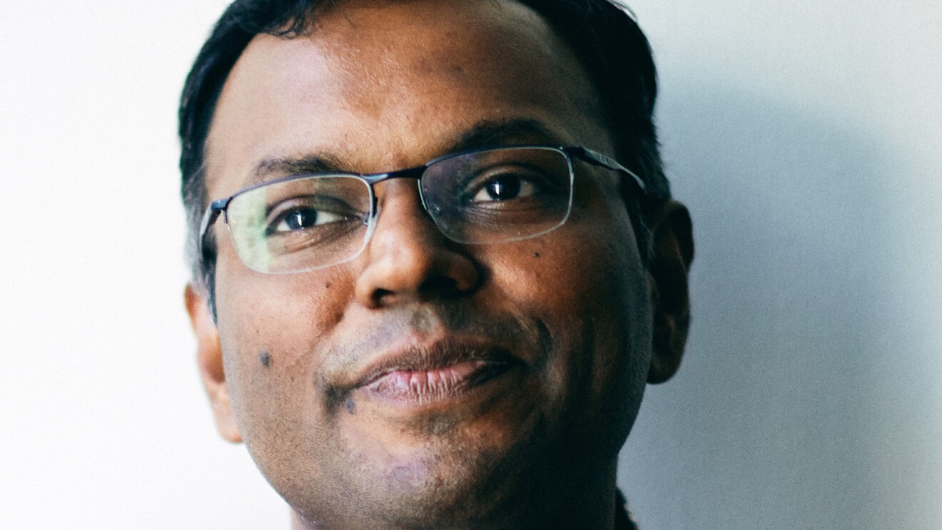 A close-cropped image of a man wearing a plaid button up over a white tee, and glasses sits in front of a white wall.