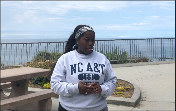 A woman wearing a sweatshirt stands on a boardwalk near a large body of water.