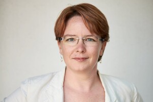 A woman smiles for a headshot photo in front of a white background while wearing a white blazer and glasses.