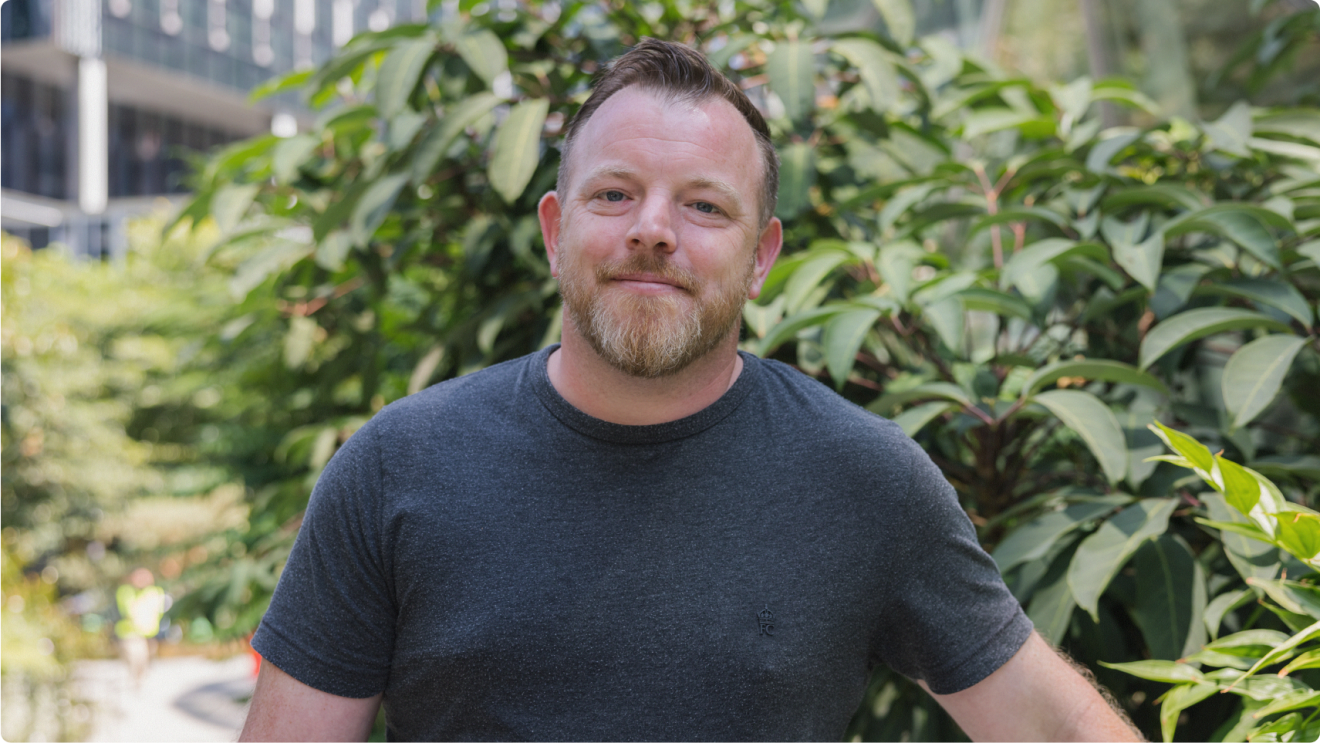 Smiling man in a dark grey t shirt standing in front of foliage