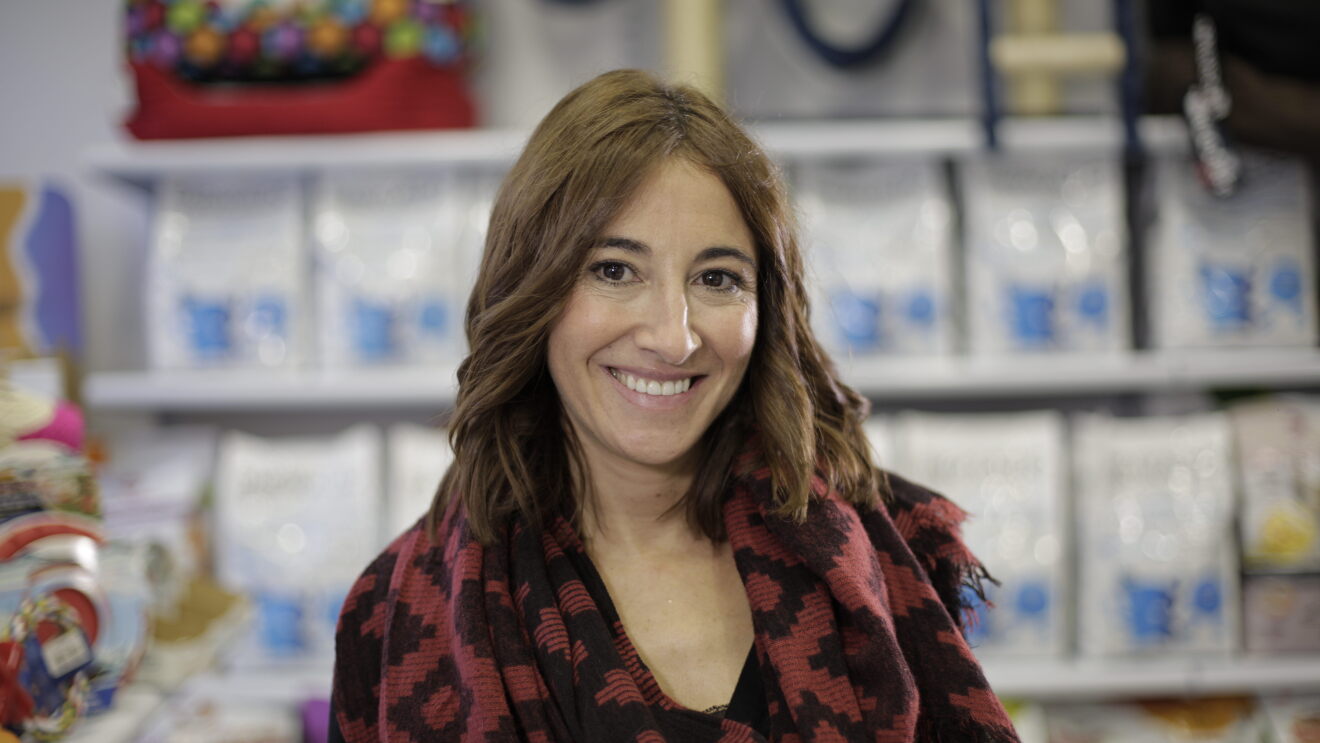 Marta Hernando de Barakaldo Vet Shop es una mujer de mediana edad con el pelo largo y con un fular de formas geométricas. En el fondo de la imagen se puede ver el pienso, uno de los productos de esta marca. 