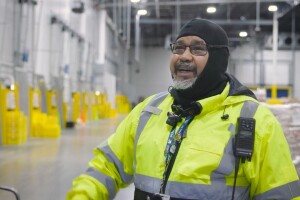 An Amazon employee smiles and wears a bright yellow safety jacket as he works in a fulfillment center.