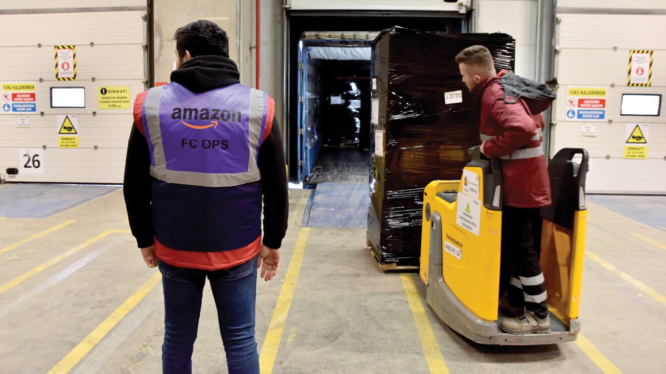 Dentro de un centro logístico, aparecen dos trabajadores de Amazon cargando un camión. La foto está dentro del centro logístico y uno de los trabajadores está de pie con un chaleco morado con el texto Amazon FC Ops. Y el otro trabajador está en un toro semiautomático de color amarillo. En el toro está cargado con un palé con material cubierto con plástico negro. El toro se está dirigiendo dentro de un camión que tiene las puertas abiertas. 