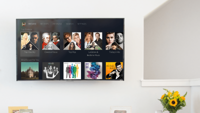 Flat screen tv on a wall showing Amazon Music displayed within Roku experience. The television is in a living room setting, flanked by flowers and personal photographs