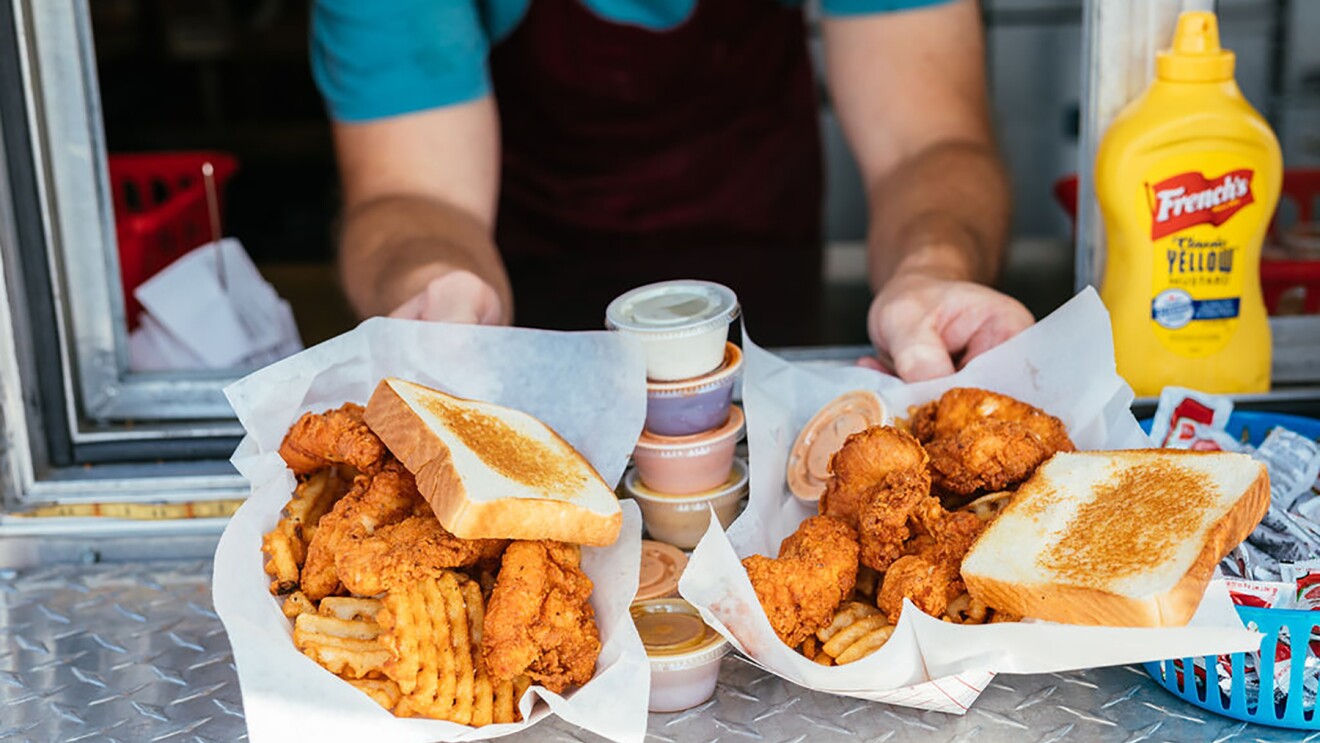 Baskets of chicken fingers, waffle fries and Texas toast are passed through the window of a food truck. A stack of condiments and sauces balances between the two baskets of food.