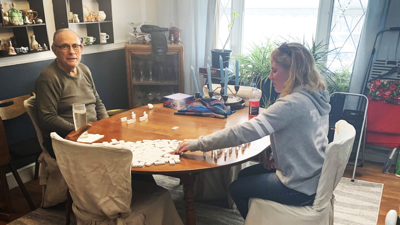 An image of a man and a woman playing dominoes at a kitchen table. The man is looking at the camera and smiling.