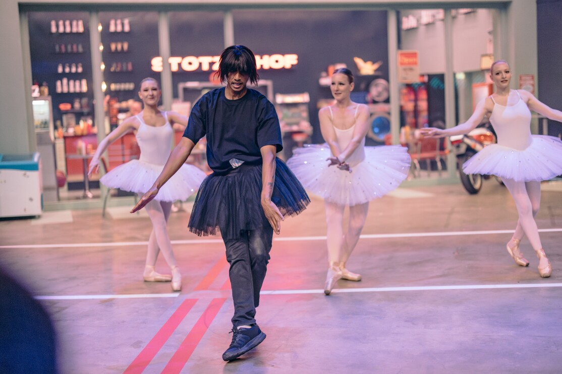 Episodenfoto von und mit Teddy Teclebrhan bei Prime Video. Man sieht Teddy in schwarzer Jeans, schwarzem T-Shirt, schwarzer Perücke und einem schwarzen Ballet-Tutu vor drei in weiß gekleideten Ballerinas tanzen.