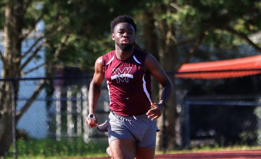 An image of a young man running on a school track wearing a track team uniform.