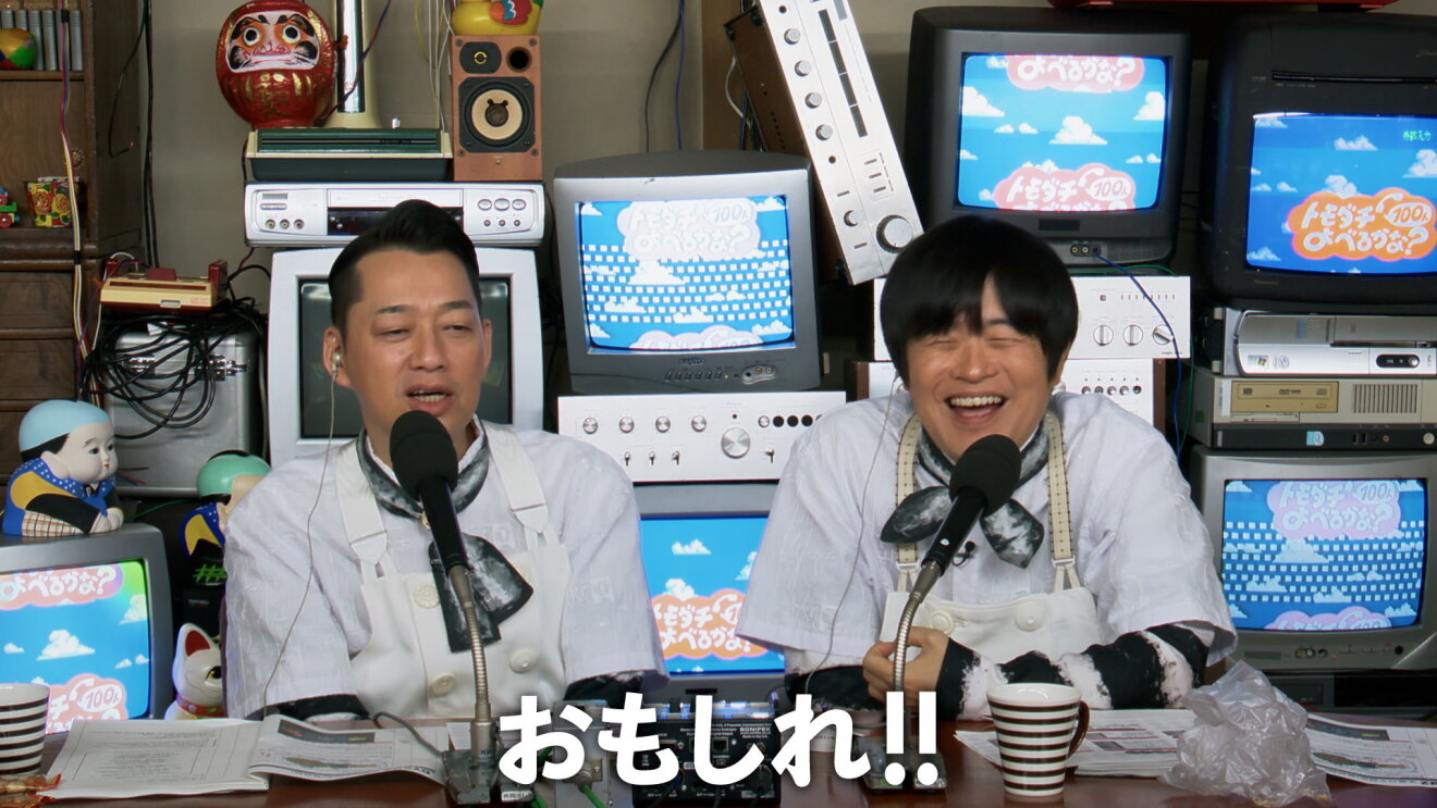 実況席のように、マイクを前に並んで座る男性2人。背後にはブラウン管テレビが多く積まれている。