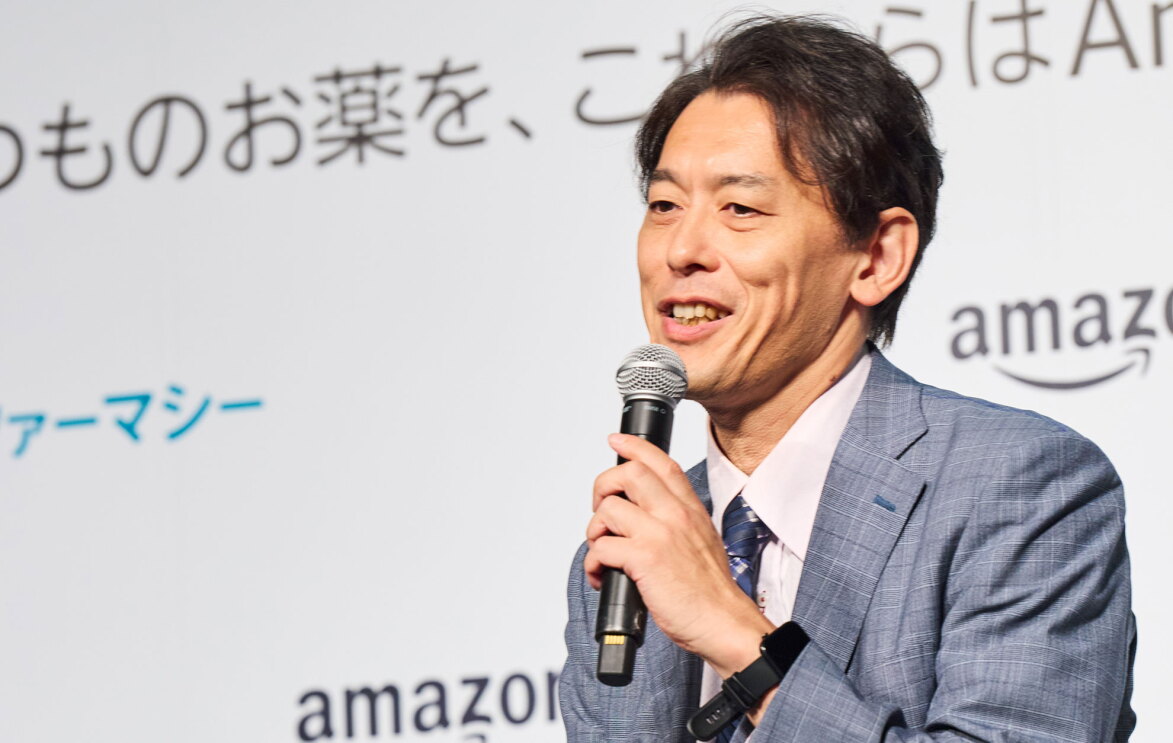 青の細かなチェック柄のスーツ姿の男性がマイクを持って話している。背景には薬の文字やAmazonのロゴが入った白いパネルがある
