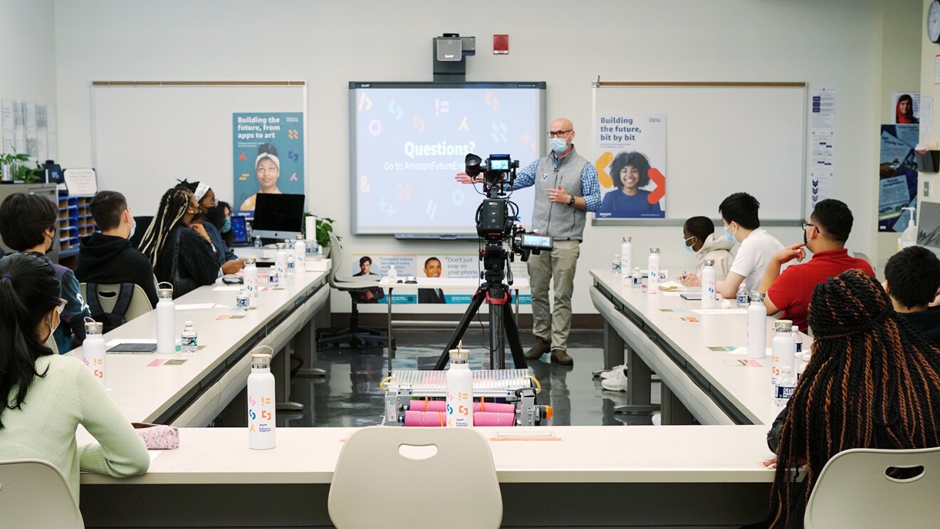 A photo of students in a classroom, sitting at a desk that surrounds a projector and faces a screen, listening to an instructor.