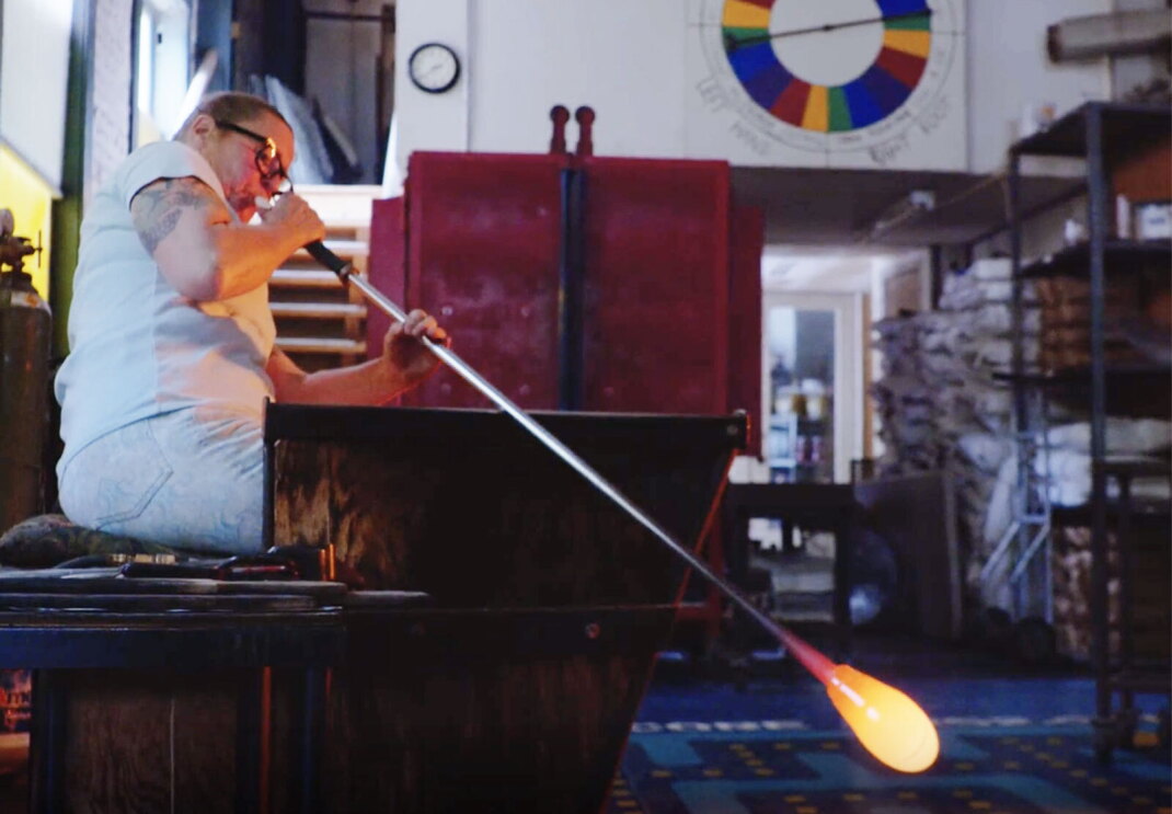 Dehanna Jones blows into a long tool to shape the glass at the end of the tool.
