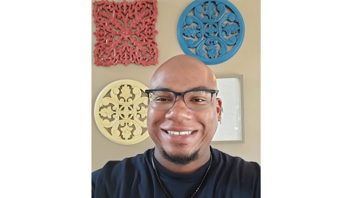 An image of a man wearing glasses and smiling for a photo at his home in front of a wall with colorful, circular decorations on it. 