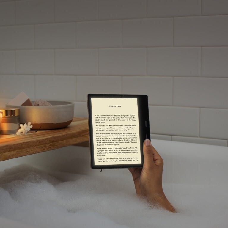 A woman holds a Kindle Oasis device with the page of a book on the display. Her arm appears from bubbles in a tub, with a wooden tub caddy behind the device.