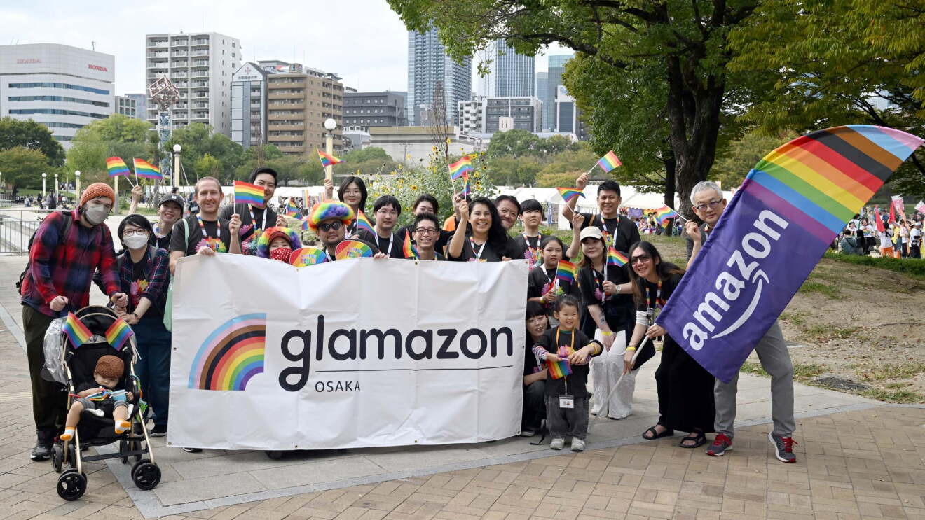 30人ぐらいの人々が「glamazon Osaka」という文字が印刷された横断幕の後ろでポーズをしている。1人の男性が大きなレインボーフラッグを持っている。