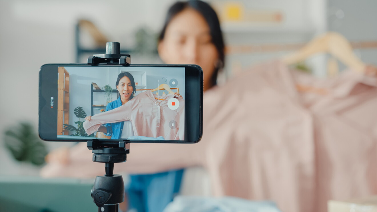 Young Asia lady fashion designer using mobile phone receiving purchase order and showing clothes recording video live streaming online at a shop.