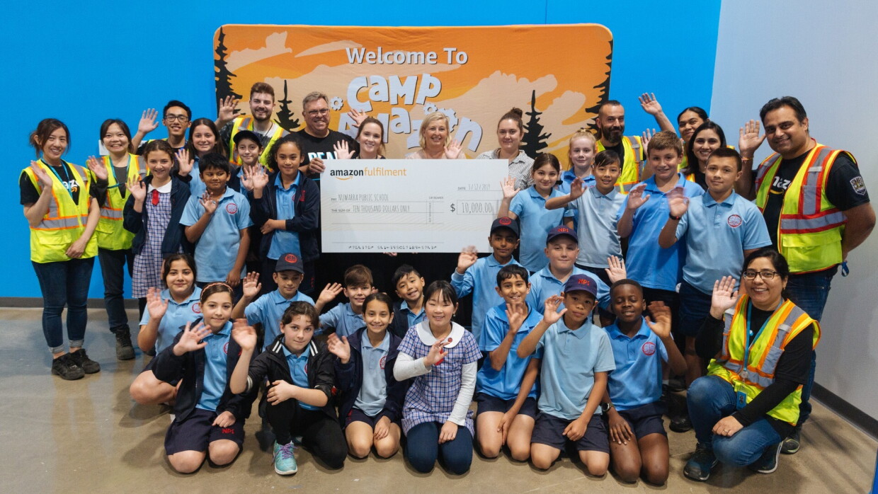 A group of kids, their teachers and Amazon Camp staff wave and smile for the camera holding a check for $10,000 from Amazon.