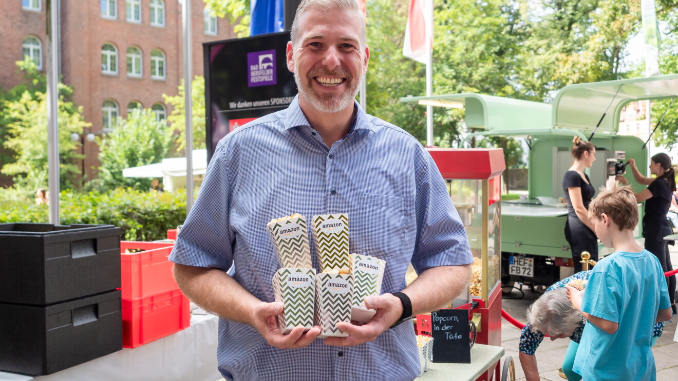 Ein Mann in Jeans und blauem Kurzarmhemd hält Popcorn-Tüten mit Amazon Aufdruck in der Hand.