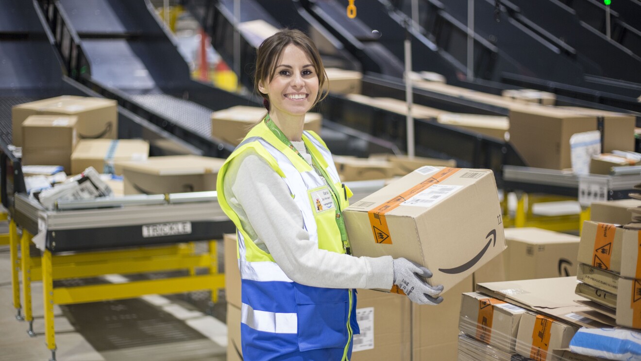 Piano americano di Ilaria di Bartolomeo, Operations Manager di Amazon a Casirate (Bergamo), mentre lavora nel centro di smistamento. Sorregge in mano un pacco con il sorriso di Amazon. 