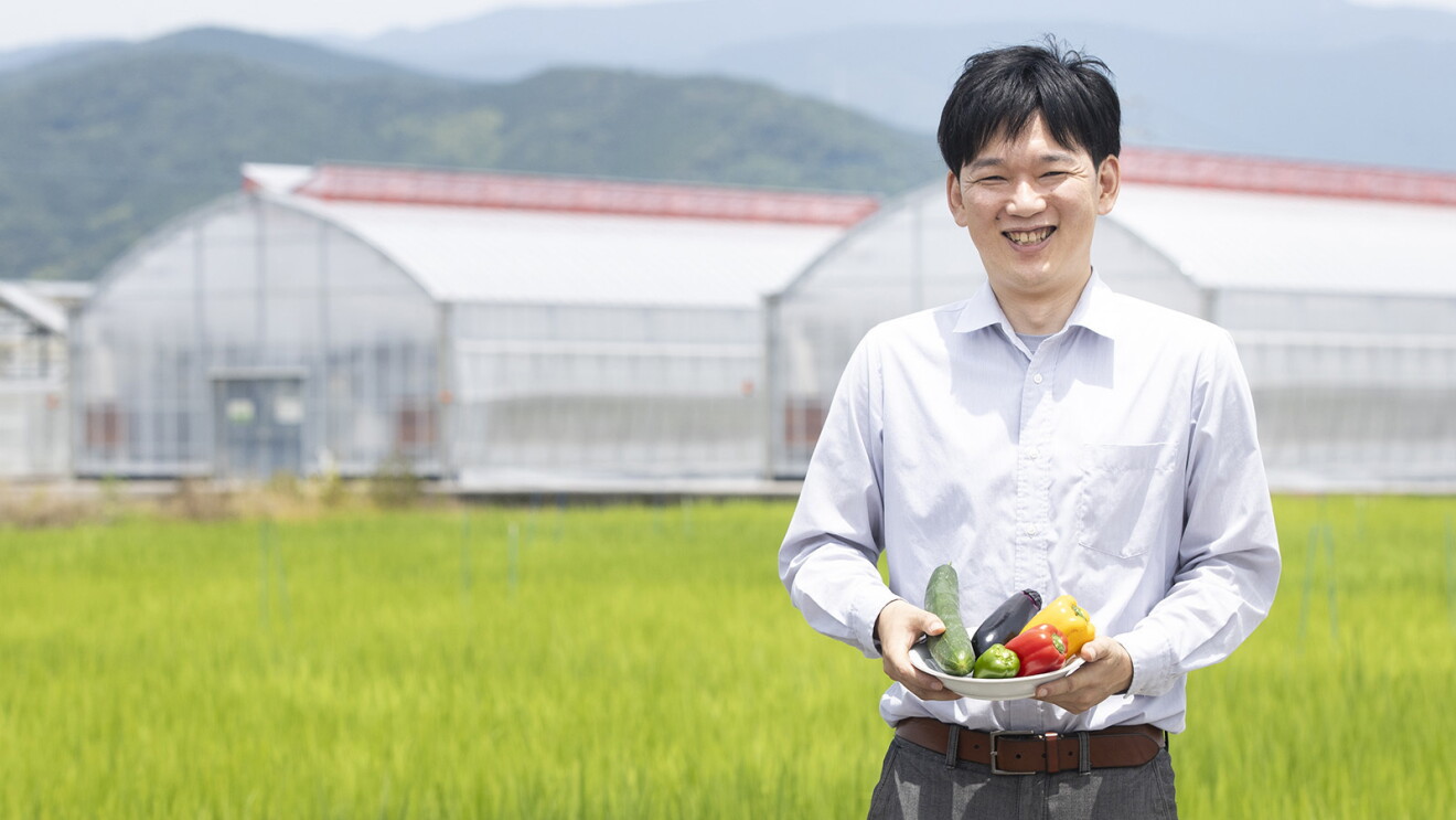 田んぼとビニールハウスの前に立ちほほ笑む男性。手には野菜