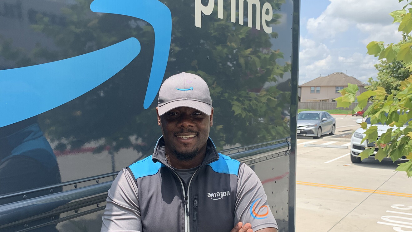An image of a man standing in front of an Amazon delivery truck with the Amazon logo and the words "Prime" written on the side. He is wearing an Amazon-branded hat and vest and smiling with his arms folded in front of him.