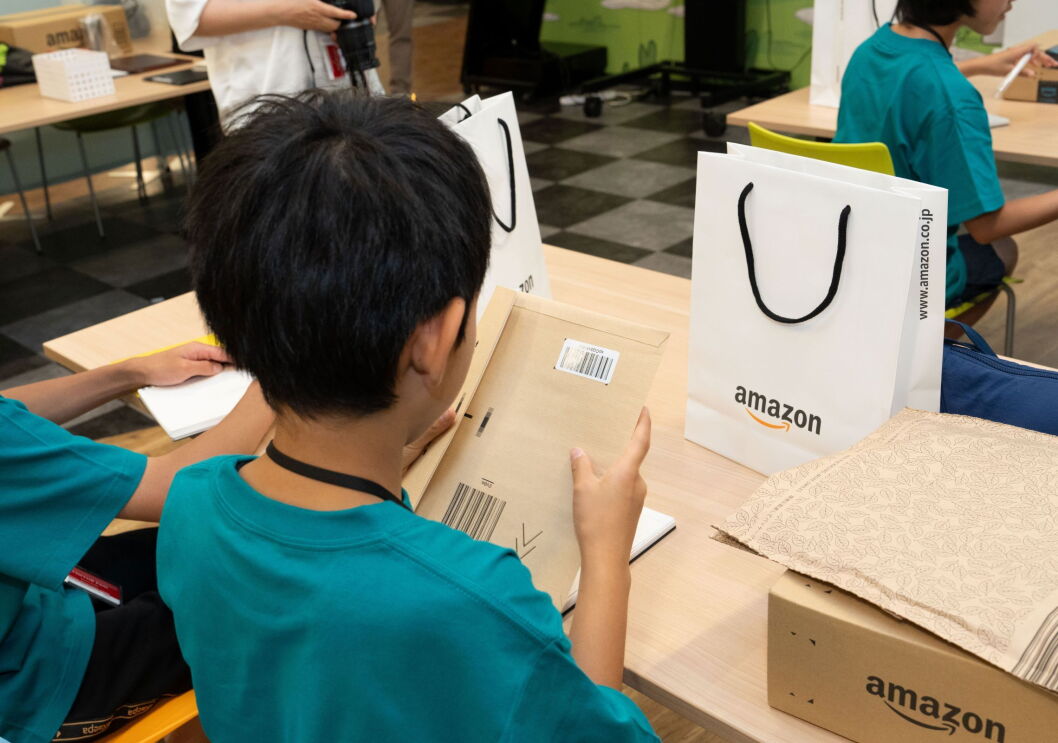 ダンボールで作られた封筒を手に持ち見ている男の子。その前にはAmazonロゴが入った白い紙袋やAmazonの段ボールが置かれている