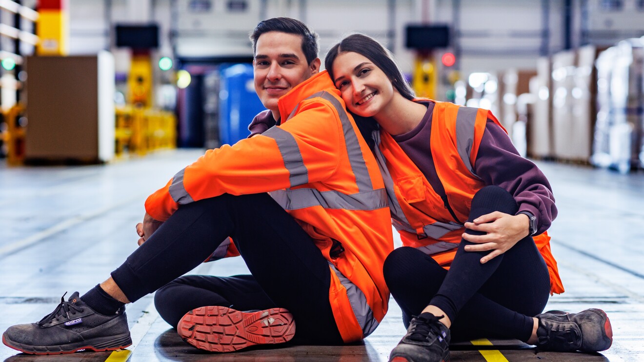 Fabiana y Rafael posando en el suelo de un centro logístico de Amazon. Los dos están posando, mirando de frente a cámara. Ella está de frente, los las piernas cruzadas, zapatos de seguridad negros, pantalón negro, camiseta morada y chaleco naranja. Lleva el suelo y es lacio y largo, Agarra con la mano derecha a Rafael. Él está de lado con las piernas cruzadas pero mirando a cámara. Lleva también zapatos de seguridad, pantalón negro, chaqueta que imita al chaleco de color naranja.